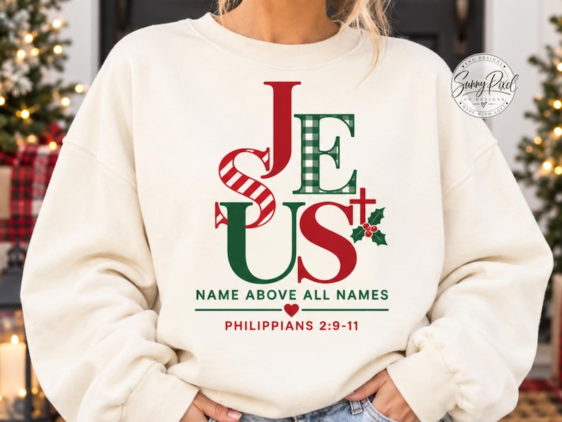 May include: A cream-colored crewneck sweatshirt featuring a graphic print that spells JESUS in red and green patterns. Below the text, it reads NAME ABOVE ALL NAMES, a small red heart, and the scripture reference PHILIPPIANS 2:9-11 in red text.