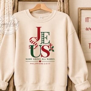 May include: A cream-colored crewneck sweatshirt featuring a graphic print of the word JESUS in red and green patterns, including stripes and plaid. Below the text, it reads NAME ABOVE ALL NAMES and PHILIPPIANS 2:9-11 with a small red heart.