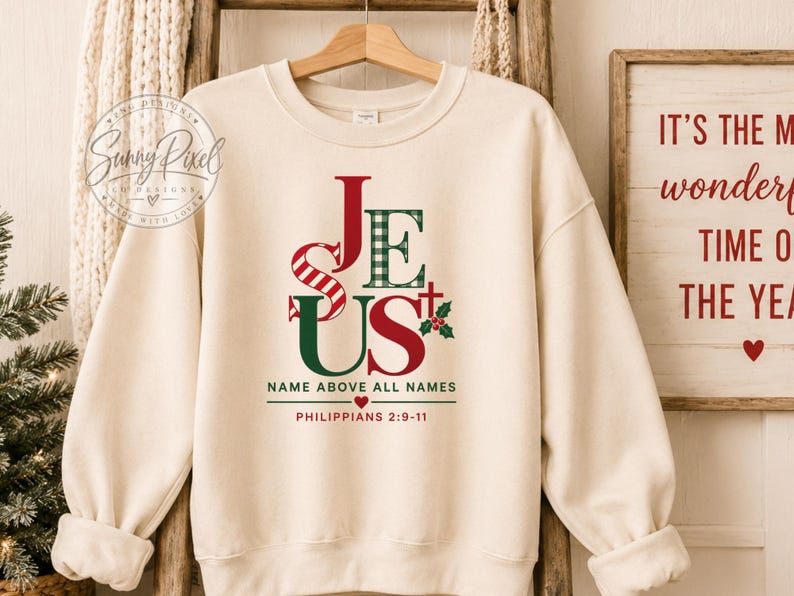 May include: A cream-colored crewneck sweatshirt featuring a graphic print of the word JESUS in red and green patterns, including stripes and plaid. Below the text, it reads NAME ABOVE ALL NAMES and PHILIPPIANS 2:9-11 with a small red heart.