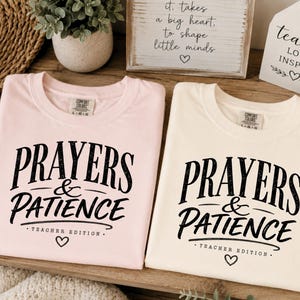 May include: Two crew neck t-shirts in light pink and cream, each featuring black screen-printed text that reads PRAYERS & PATIENCE TEACHER EDITION with a small heart icon below. The shirts have a Comfort Colors brand label at the inner neckline.