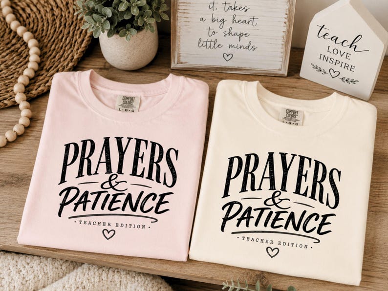 May include: Two crew neck t-shirts in light pink and cream, each featuring black screen-printed text that reads PRAYERS & PATIENCE TEACHER EDITION with a small heart icon below. The shirts have a Comfort Colors brand label at the inner neckline.