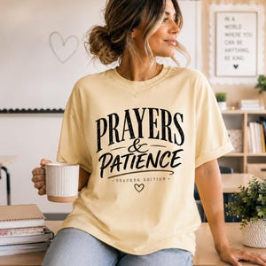 May include: A light yellow short-sleeve t-shirt featuring black text that reads PRAYERS & PATIENCE TEACHER EDITION with a small heart icon below the lettering. The shirt has a crew neckline and a relaxed fit, made of a solid-colored fabric.