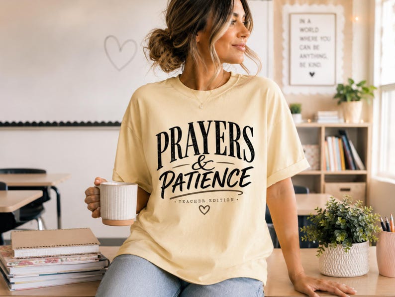 May include: A light yellow short-sleeve t-shirt featuring black text that reads PRAYERS & PATIENCE TEACHER EDITION with a small heart icon below the lettering. The shirt has a crew neckline and a relaxed fit, made of a solid-colored fabric.