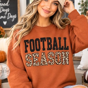 May include: A rust-colored long-sleeve sweatshirt featuring the text FOOTBALL SEASON printed on the front. The word FOOTBALL is in solid black block letters, while the word SEASON is in a leopard print pattern with a black outline.