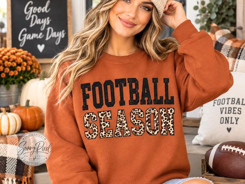May include: A rust-colored long-sleeve sweatshirt featuring the text FOOTBALL SEASON printed on the front. The word FOOTBALL is in solid black block letters, while the word SEASON is in a leopard print pattern with a black outline.