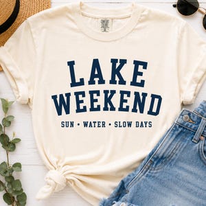 K&ouml;nnte beinhalten: Ein cremefarbenes Kurzarm-T-Shirt mit dem dunkelblauen Schriftzug LAKE WEEKEND in einer fetten, gebogenen Schriftart, wobei die W&ouml;rter SUN &bull; WATER &bull; SLOW DAYS in einer kleineren, geraden Schriftart unter dem Haupttext auf der Vorderseite stehen.
