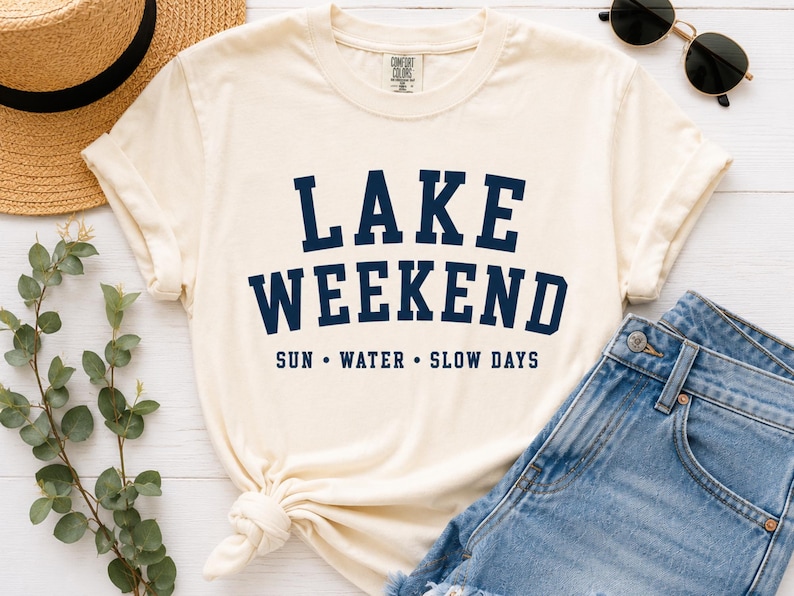 K&ouml;nnte beinhalten: Ein cremefarbenes Kurzarm-T-Shirt mit dem dunkelblauen Schriftzug LAKE WEEKEND in einer fetten, gebogenen Schriftart, wobei die W&ouml;rter SUN &bull; WATER &bull; SLOW DAYS in einer kleineren, geraden Schriftart unter dem Haupttext auf der Vorderseite stehen.