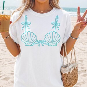 May include: White t-shirt featuring a turquoise seashell bra design. The graphic includes two seashell cups with bows and straps. The shirt is a casual, beach-themed apparel item.