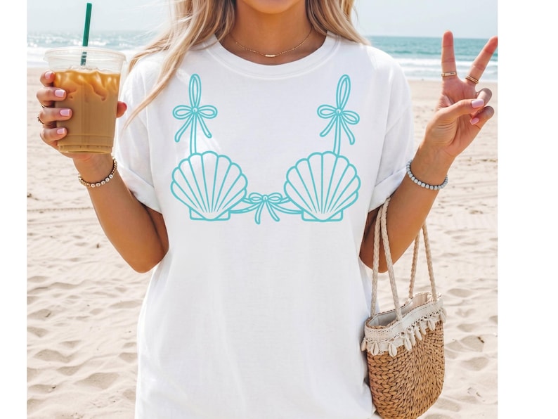 May include: White t-shirt featuring a turquoise seashell bra design. The graphic includes two seashell cups with bows and straps. The shirt is a casual, beach-themed apparel item.
