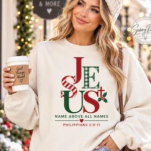 May include: A cream-colored crewneck sweatshirt featuring the word JESUS in red and green plaid lettering. Below the text, it reads NAME ABOVE ALL NAMES, a small heart, and PHILIPPIANS 2:9-11. A small red cross and green holly leaves are visible.