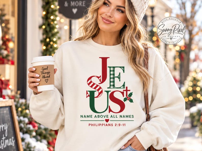 May include: A cream-colored crewneck sweatshirt featuring the word JESUS in red and green plaid lettering. Below the text, it reads NAME ABOVE ALL NAMES, a small heart, and PHILIPPIANS 2:9-11. A small red cross and green holly leaves are visible.