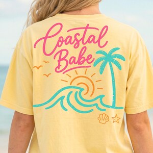 May include: Yellow t-shirt with a beach-themed graphic on the back. The design includes the words "Coastal Babe" in pink and a turquoise palm tree, waves, and sun. A seashell and star are also included.
