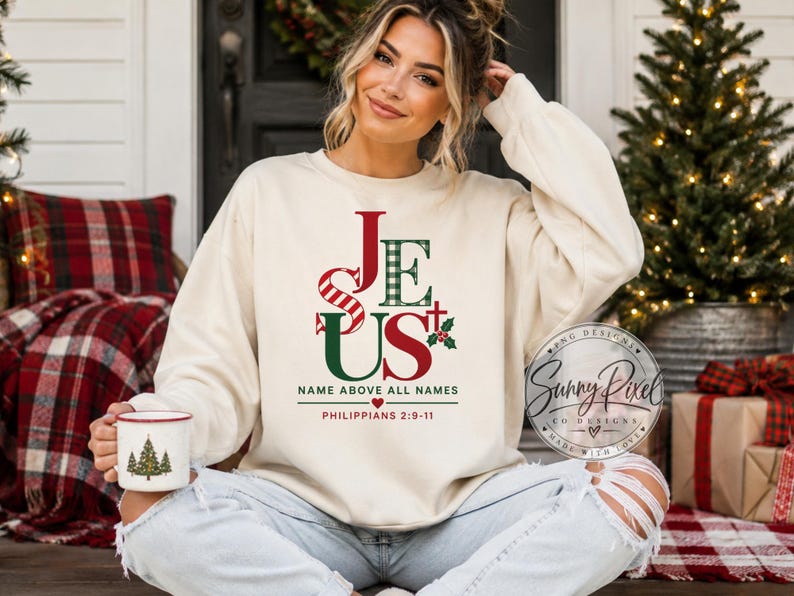 May include: A cream-colored crewneck sweatshirt featuring a graphic print that reads JESUS in red and green, with the text NAME ABOVE ALL NAMES and PHILIPPIANS 2:9-11 below. The design includes a small cross, holly leaves, and a red heart.