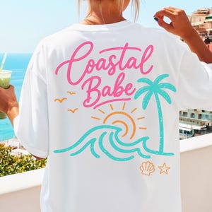 May include: White t-shirt with a colorful coastal design. The back of the shirt features the words "Coastal Babe" in pink script, along with a palm tree, sun, waves, birds, a seashell, and a star. The shirt is a relaxed fit.