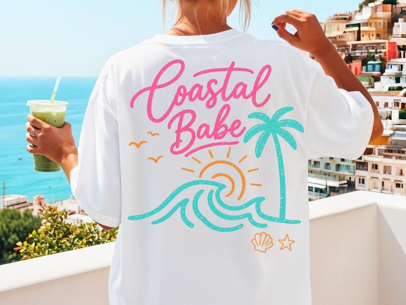 May include: White t-shirt with a colorful coastal design. The back of the shirt features the words "Coastal Babe" in pink script, along with a palm tree, sun, waves, birds, a seashell, and a star. The shirt is a relaxed fit.