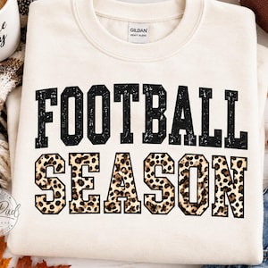 May include: A cream-colored crewneck sweatshirt featuring the text FOOTBALL SEASON in a bold, block-letter font. The word FOOTBALL is printed in a distressed black texture, while the word SEASON features a brown and black leopard print pattern.