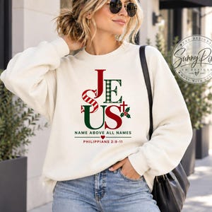 May include: A cream-colored crewneck sweatshirt featuring the word JESUS in red and green plaid lettering, with a small cross and holly sprig. Below the text, it reads NAME ABOVE ALL NAMES and PHILIPPIANS 2:9-11 with a small red heart icon.