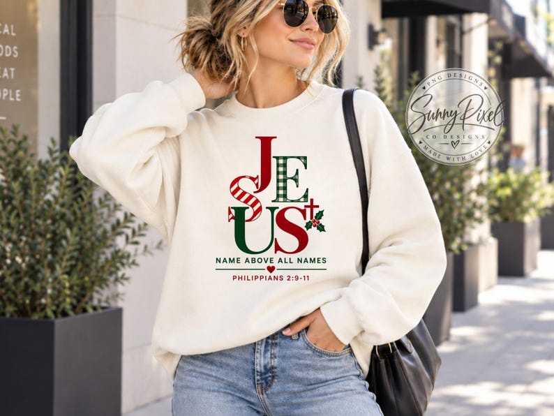 May include: A cream-colored crewneck sweatshirt featuring the word JESUS in red and green plaid lettering, with a small cross and holly sprig. Below the text, it reads NAME ABOVE ALL NAMES and PHILIPPIANS 2:9-11 with a small red heart icon.