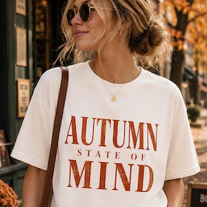 Op de afbeelding: Een crèmekleurig T-shirt met korte mouwen en de tekst "AUTUMN STATE OF MIND" in een verweerd, roestoranje lettertype op de borst. Het shirt heeft een ronde hals en een relaxte pasvorm, met een bruine band zichtbaar over de schouder.