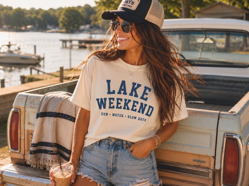 K&ouml;nnte beinhalten: Ein cremefarbenes Kurzarm-T-Shirt mit dem dunkelblauen Schriftzug LAKE WEEKEND in fetter Schrift und dem kleineren Text SUN WATER SLOW DAYS darunter. Das T-Shirt hat einen Rundhalsausschnitt und eine lockere Passform aus baumwollartigem Stoff.