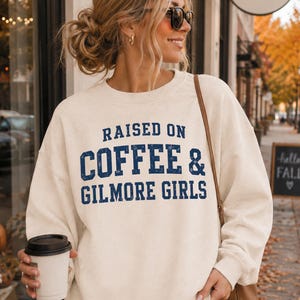 May include: A cream-coloured crewneck sweatshirt featuring the dark blue distressed text RAISED ON COFFEE & GILMORE GIRLS printed across the chest. The long-sleeved garment has a ribbed neckline, cuffs, and hem, providing a relaxed fit.