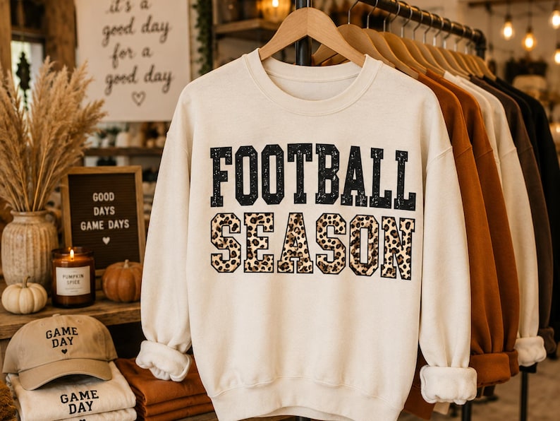 May include: A cream-colored crewneck sweatshirt featuring the text FOOTBALL SEASON printed on the front. The word FOOTBALL is in black, and the word SEASON is in a brown and black leopard print pattern. The sweatshirt has long sleeves and a ribbed collar.
