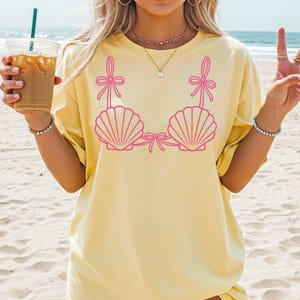 May include: A light yellow t-shirt featuring a pink seashell bra design. The graphic includes two seashell cups with bows and straps. The shirt is worn on a beach setting.