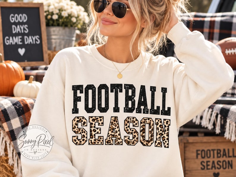 May include: A cream-colored crewneck sweatshirt featuring the text FOOTBALL SEASON printed on the front. The word FOOTBALL is in a distressed black font, while the word SEASON is in a black-outlined font filled with a brown leopard print pattern.