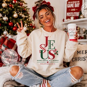 May include: A cream-colored crewneck sweatshirt featuring the word JESUS in red and green, with the text NAME ABOVE ALL NAMES and PHILIPPIANS 2:9-11 printed below. The design includes a small cross and holly leaves, paired with light blue distressed jeans.