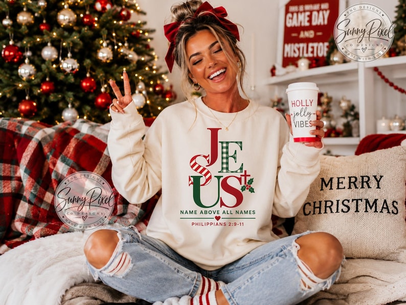 May include: A cream-colored crewneck sweatshirt featuring the word JESUS in red and green, with the text NAME ABOVE ALL NAMES and PHILIPPIANS 2:9-11 printed below. The design includes a small cross and holly leaves, paired with light blue distressed jeans.