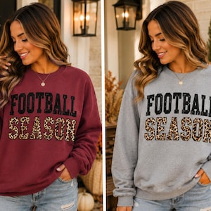 May include: A long-sleeve crewneck sweatshirt in maroon or heather grey featuring the text FOOTBALL SEASON printed on the front. The word FOOTBALL is in black block letters, and the word SEASON is in a brown and black leopard print pattern.