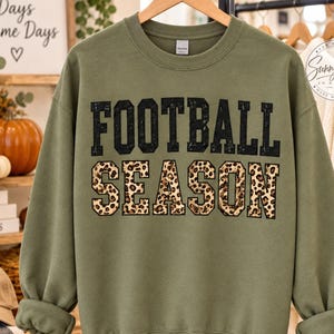 May include: An olive green crewneck sweatshirt featuring the text FOOTBALL SEASON in a bold, block-letter font. The word FOOTBALL is printed in solid black, while the word SEASON is filled with a brown and black leopard print pattern.