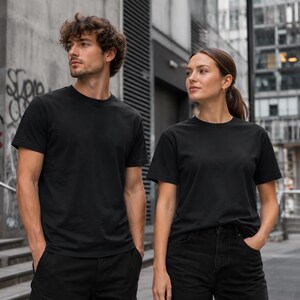 Könnte beinhalten: Zwei Personen tragen draußen schwarze T-Shirts mit Rundhalsausschnitt und schwarze Hosen. Die T-Shirts sind schlicht und haben kurze Ärmel. Der Hintergrund zeigt ein Gebäude und eine Wand mit Graffiti.