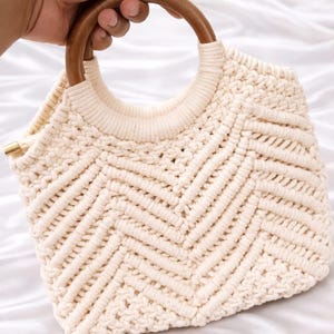 May include: Cream-colored macrame handbag with a geometric chevron pattern. The bag features a rounded wooden handle and a structured design. The bag is held by a hand, showcasing the natural texture and craftsmanship of the woven material.
