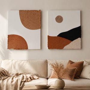 May include: Two abstract wall art pieces. One features a brown half-circle and a smaller brown circle on a white background. The other has a black mountain shape, a brown and beige landscape, and a circular sun on a white background.
