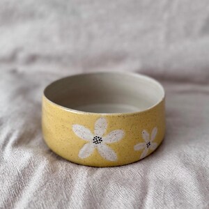 May include: A yellow ceramic bowl with a speckled finish and two white daisy flower designs. The interior of the bowl is a light grey colour. The bowl is a round shape.