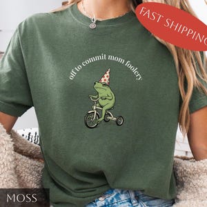 May include: A moss green t-shirt with a cartoon frog on a tricycle, wearing a party hat. The text "off to commit mom foolery" is arched above the frog. A red oval with "FAST SHIPPING" is in the upper right corner.
