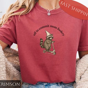 Op de afbeelding: Karmizijnrood T-shirt met een illustratie van een wasbeer op een scooter met een tovenaars hoed. De tekst luidt "off to commit mom foolery". Een leuke, grillige illustratie.