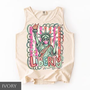 Könnte beinhalten: Ein elfenbeinfarbenes Tanktop mit einer Grafik der Freiheitsstatue, die eine Sonnenbrille trägt und eine rosa Kaugummiblase macht. Das Design zeigt eine Fackel, ein Friedenszeichen, Sterne, Blitze und das Wort LIBERTY in bunter Schrift.