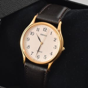 May include: A gold-tone wristwatch with a cream-colored dial, black Arabic numerals, and thin hands, featuring a dark brown leather strap. The watch is presented inside a black textured box with a matching lid resting at an angle.