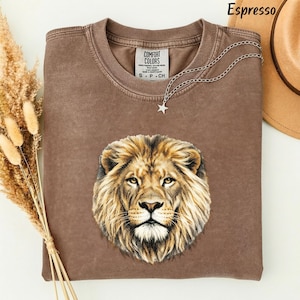 May include: A brown crew neck t-shirt featuring a detailed graphic of a lion's face centered on the chest. The shirt has a tag that reads Comfort Colors, and a silver-toned chain necklace with a small star charm rests on the collar.