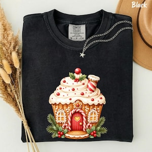Può includere: Una t-shirt nera a girocollo con la grafica di una casetta di pan di zenzero a forma di cupcake, con glassa bianca, bacche rosse e un camino a bastoncino di zucchero. La maglia presenta un'etichetta Comfort Colors e una catenina con stella.