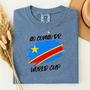 May include: A blue t-shirt with the text "GO CONGO DR WORLD CUP" and a graphic of the Democratic Republic of the Congo flag. A silver necklace with a star pendant is draped on the shirt. A brown hat is visible in the background.
