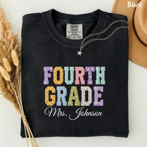 Puede incluir: Una camiseta negra de cuello redondo con el texto FOURTH GRADE en letras grandes y coloridas con varios patrones, incluyendo cuadros vichy, lunares y flores. Debajo del texto, el nombre Mrs. Johnson está escrito en letra cursiva blanca.