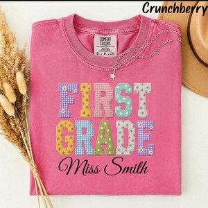 以下が含まれることがあります： ピンクの半袖Tシャツ。花柄やギンガムチェックなど、様々な模様が施されたカラフルなブロック体の文字で「FIRST GRADE」とプリントされています。その下には、黒い筆記体フォントで「Miss Smith」という名前が記されています。