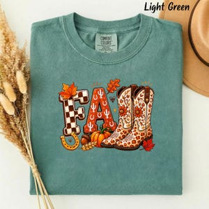 May include: A light green t-shirt featuring a graphic print of the word FALL. The letter F is checkered, the A is orange with white cactus icons, and the L is replaced by two floral-patterned cowboy boots. Includes a pumpkin and leaves.