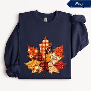 Puede incluir: Una sudadera azul marino de cuello redondo con un gran gráfico central de una hoja de arce compuesta por varios patrones de patchwork en tonos naranja, rojo, amarillo y marrón, incluyendo cuadros, vichy y estampados florales.