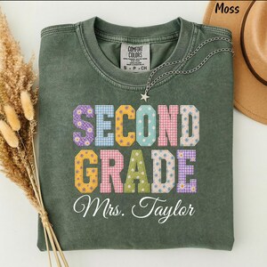 Puede incluir: Una camiseta de cuello redondo color verde musgo con el texto SECOND GRADE en letras grandes y coloridas con varios patrones, incluyendo cuadros vichy, lunares y margaritas. Debajo, el texto Mrs. Taylor está impreso en cursiva blanca.