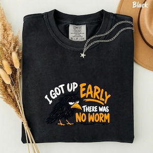 May include: A black short-sleeved t-shirt featuring a graphic of a grumpy-looking black bird with messy feathers and a yellow beak. The text reads I GOT UP EARLY THERE WAS NO WORM in white and yellow lettering. The shirt has a Comfort Colors label.