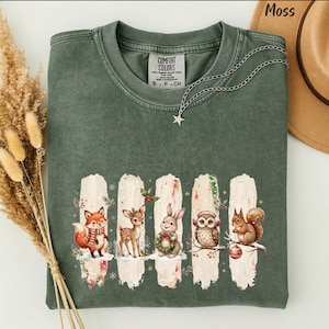 May include: A moss green crewneck t-shirt with a Comfort Colors label at the neck. The front features five vertical white brushstroke graphics, each containing a woodland animal: a fox, a deer, a rabbit, an owl, and a squirrel, all with winter accents.
